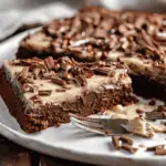 A rich Chocolate Texas Sheet Cake topped with creamy buttermilk frosting and chocolate shavings on a white plate.