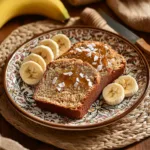 Two slices of banana bread topped with coconut flakes and jam, served on a decorative plate with sliced bananas on the side.