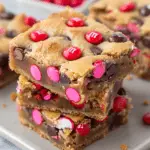 A stack of Valentine's Day M&M Cookie Bars loaded with red, pink, and chocolate candies on a plate.