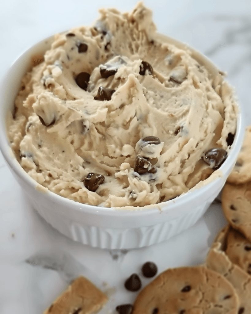 A creamy bowl of cookie dough dip with chocolate chips, surrounded by chocolate chip cookies on a marble surface.