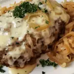 A juicy crockpot French onion meatloaf topped with melted Swiss cheese, caramelized onions, and fresh parsley, served with sautéed onions on a white plate.