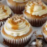 A plate of salted caramel cheesecake cupcakes topped with creamy frosting, caramel drizzle, and chocolate shavings, with a spoonful of cake beside them.
