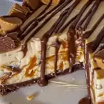 Creamy Reese’s peanut butter cup cheesecake with a chocolate crust, swirls of caramel, and drizzled chocolate, topped with peanut butter cups.