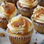 A close-up of salted caramel cheesecake cupcakes topped with creamy frosting, drizzled with caramel sauce, and sprinkled with chocolate shavings.