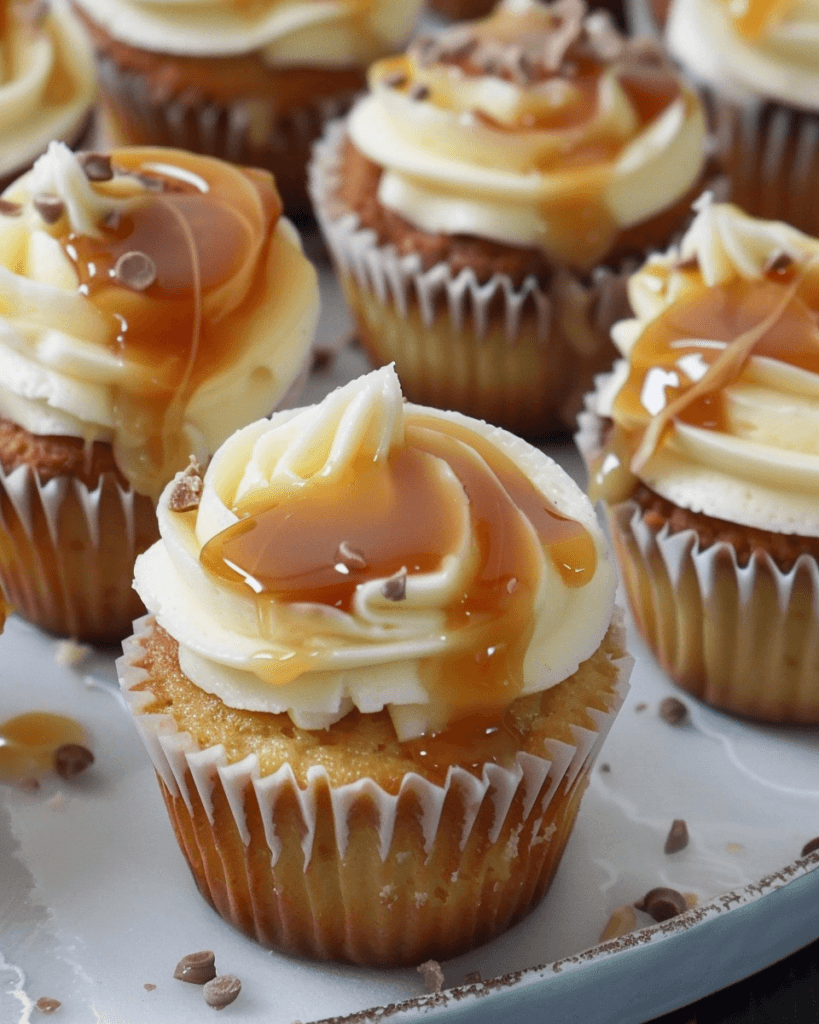 A close-up of salted caramel cheesecake cupcakes topped with creamy frosting, drizzled with caramel sauce, and sprinkled with chocolate shavings.