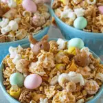 A festive Easter Bunny Bait snack mix with cereal, pretzels, pastel chocolate eggs, bunny-shaped treats, and white chocolate drizzle in blue bowls.