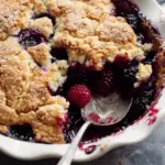 A close-up of a freshly baked mixed berry cobbler with a golden, flaky crust and juicy raspberries and blackberries, with a spoon scooping out a portion.