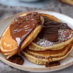 A stack of fluffy pancakes covered in rich, melted Nutella and dusted with powdered sugar, served on a plate with chocolate drizzles around.