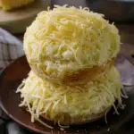 Two soft and buttery Filipino ensaymadas stacked on a wooden plate, generously topped with creamy butter and shredded cheese.