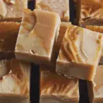 Squares of creamy caramel apple fudge with swirls of caramel on top, stacked on a dark surface.