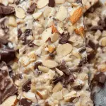 A close-up of a piece of chocolate almond bark topped with shredded coconut, almond slices, and chunks of chocolate.