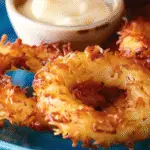 Golden-brown coconut-crusted pineapple rings served on a blue plate with a small bowl of creamy dipping sauce.