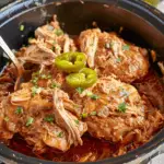 Slow-cooked shredded chicken in a savory brown sauce, topped with sliced jalapeños and chopped herbs, served in a crock pot.