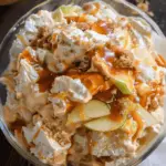 A glass bowl filled with creamy caramel apple fluff salad, featuring chunks of cheesecake filling, green apple slices, crushed graham crackers, and rich caramel drizzle.