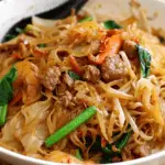 Bowl of stir-fried rice noodles with vegetables, ground meat, shrimp, and soy-based sauce, garnished with green onions and bean sprouts.