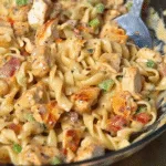 Creamy pasta with rotini noodles, grilled chicken pieces, diced bell peppers, and herbs in a savory sauce served in a bowl with a spoon.