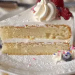 A close-up of a slice of fluffy vanilla cake with white frosting, topped with raspberries, sprinkles, and whipped cream.