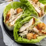 Three lettuce wraps filled with grilled chicken slices, Caesar dressing, croutons, and shaved Parmesan, served on a black plate with parchment paper.