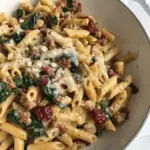 A hearty bowl of penne pasta mixed with ground sausage, wilted spinach, sun-dried tomatoes, and melted cheese.