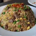 A dome-shaped serving of spicy brisket fried rice with peas, scrambled egg, scallions, and diced brisket, topped with chili sauce on a white plate.