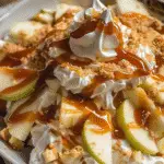 Bowl of caramel apple fluff salad topped with whipped cream peaks, green apple slices, caramel drizzle, and crumbled cookie bits.