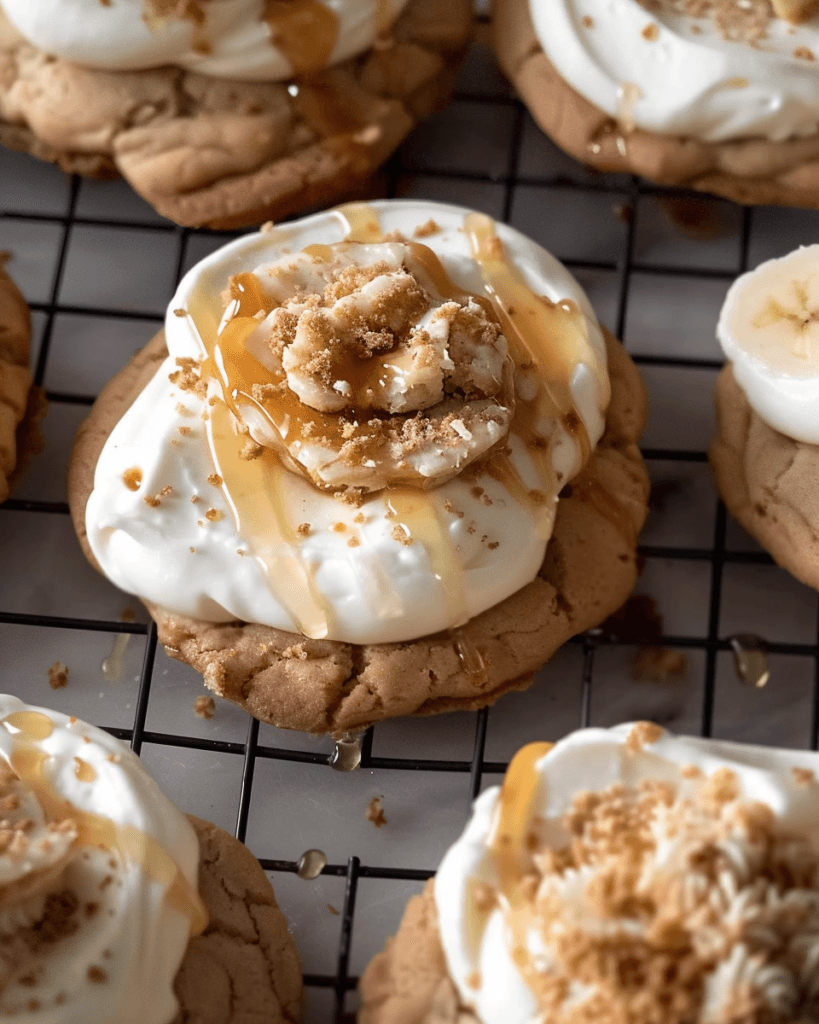 Close-up of a banoffee pie cookie topped with whipped cream, banana slices, caramel drizzle, and cookie crumbles, cooling on a wire rack.