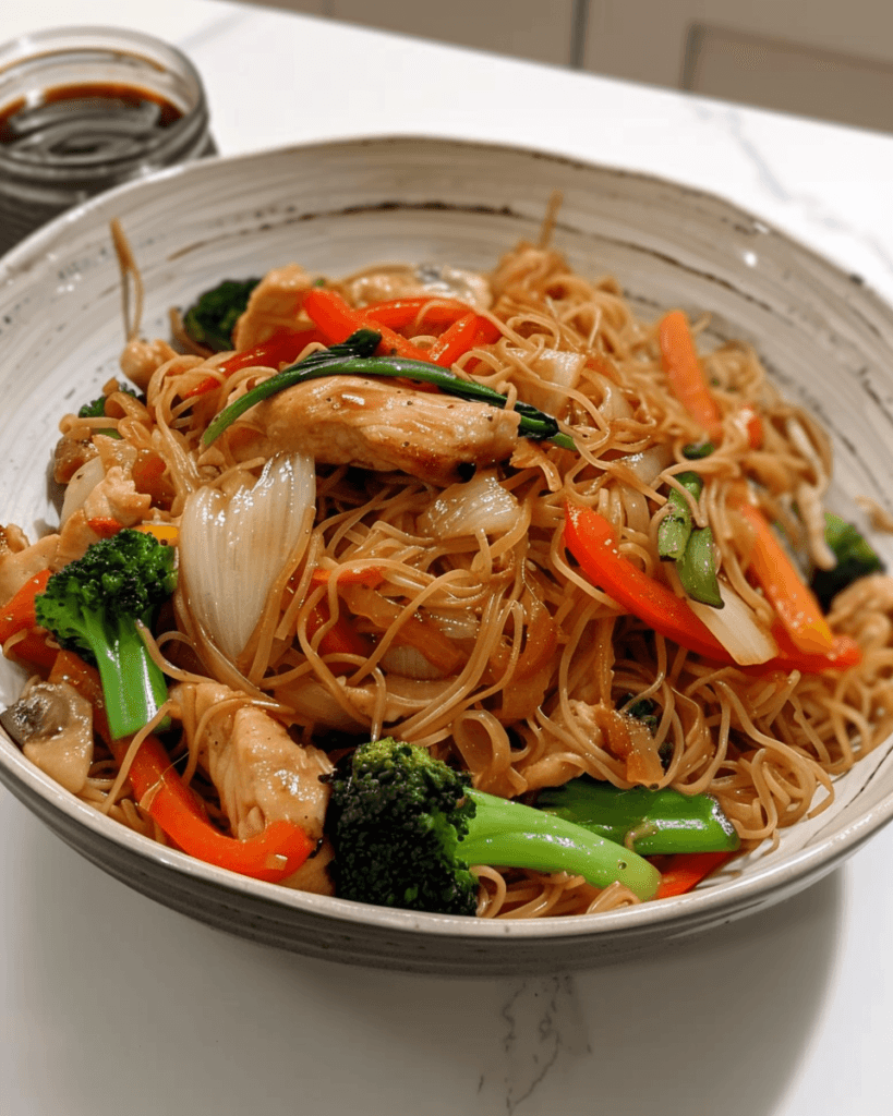 Bowl of stir-fried thin rice noodles with chicken, broccoli, red bell peppers, carrots, onions, and a savory soy-based sauce.
