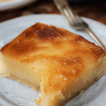 A close-up of a golden-brown slice of cassava cake with a dense, moist texture and a glossy top, served on a plate with a fork.