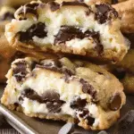 Close-up of a cheesecake-stuffed chocolate chip cookie cut in half, revealing a creamy cheesecake filling surrounded by gooey melted chocolate and golden-brown cookie dough.