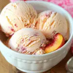 Three scoops of homemade peach ice cream with peach swirl in a white bowl, garnished with a fresh peach slice.