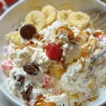Banana split fluff salad with whipped cream, banana slices, raspberries, chocolate candies, caramel drizzle, and crushed graham crackers in a white bowl.