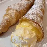 Close-up of two flaky cannoli shells dusted with powdered sugar, one filled with creamy pastry custard and served with a scoop of vanilla ice cream.