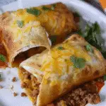 Two golden-brown beef and cheese chimichangas on a white plate, one cut open to show the seasoned meat filling, topped with melted shredded cheese and fresh cilantro.