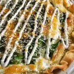 Close-up of a golden-brown cabbage pie topped with melted cheese, wilted greens, sesame seeds, and a drizzle of dill cream sauce.