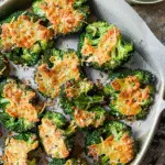 Baked smashed broccoli florets topped with melted, crispy cheese and seasoned with salt and pepper on a parchment-lined tray.