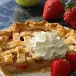 Slice of lattice-topped fruit pie with visible apple and rhubarb filling, topped with whipped cream and served with fresh strawberries.