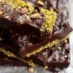 Stack of thick chocolate halva bars with a golden halva base and dark chocolate topping, sprinkled with crushed pistachios on parchment paper.