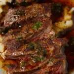 Juicy slices of marinated steak topped with fresh herbs and a rich, glossy sauce, served over creamy mashed potatoes.