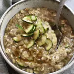 Bowl of creamy oatmeal with visible oats, topped with sautéed zucchini slices and a drizzle of syrup.