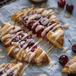 Close-up of golden-brown cherry turnovers drizzled with vanilla glaze and dusted with powdered sugar, surrounded by fresh cherries on parchment paper.