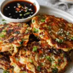 A plate of crispy, pan-fried vegetable pancakes garnished with chopped scallions and sesame seeds, served with a dark Asian dipping sauce.