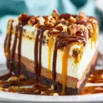Close-up of a slice of layered toffee crunch cheesecake topped with chopped pecans, chocolate, and caramel sauce.