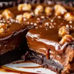 A rich slice of caramel brownie pie topped with glossy caramel sauce, chopped nuts, and sea salt.