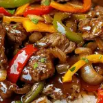 Close-up of Chinese pepper steak served over white rice, featuring tender beef strips, sautéed bell peppers in red, yellow, and green, onions, and a glossy brown stir-fry sauce.