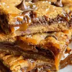 Stack of gooey salted caramel chocolate chip cookie bars with layers of melted chocolate, rich caramel, and golden cookie dough, topped with sea salt flakes.